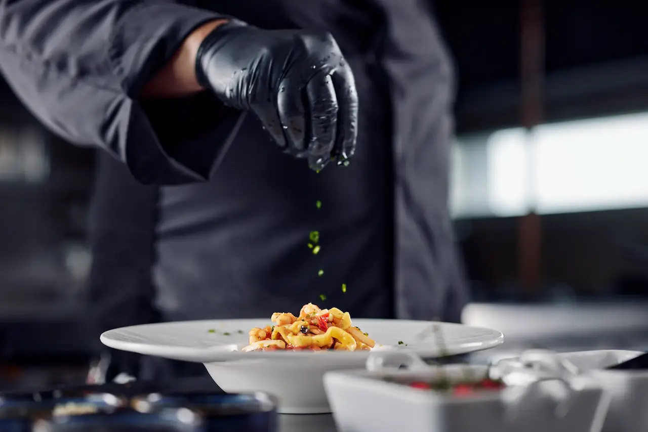 Un cuoco, in veste professionale e con un guanto di lattice, guarnisce un piatto di pasta